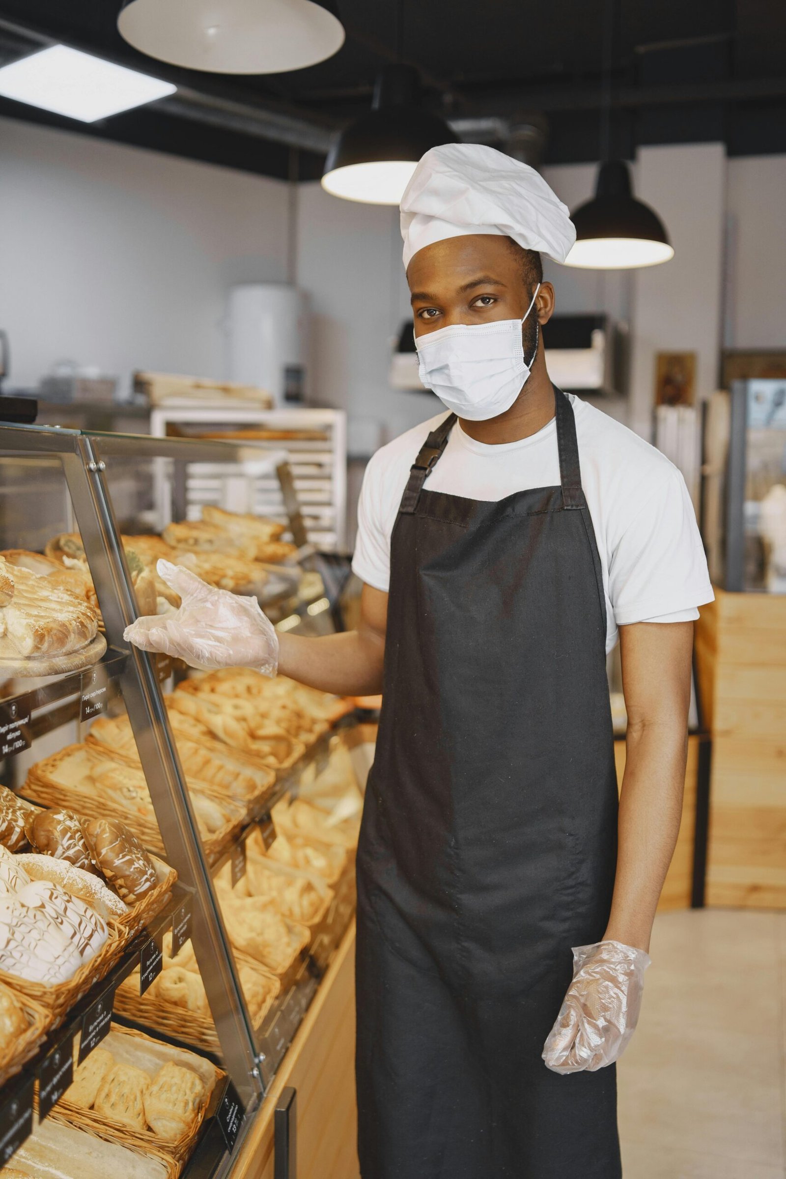 Inicio Masked baker presenting an assortment of fresh pastries in a bakery. Indoor shot.